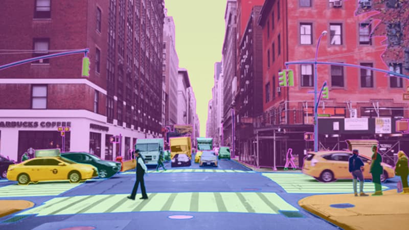A city street which includes tall buildings, cars, streetlights and pedestrians. Each object is colored differently to represent instance segmentation for autonomous vehicles.
