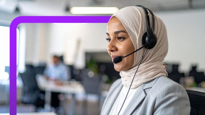 The image shows a close-up of a woman in an office setting. She is wearing a light-colored hijab and a light gray blazer. She has a black headset with a microphone. She is smiling slightly and appears to be speaking or listening to someone. In the background, the office environment is blurred, and another person is visible seated at a desk. A thick purple line frames the top and left edges of the image.