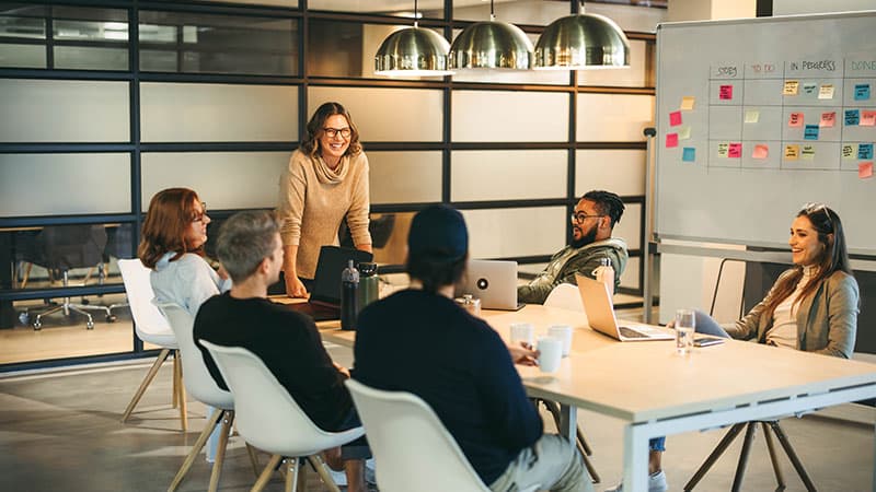 A woman stands at the head of a meeting table while her colleagues sit and listen to her talk.