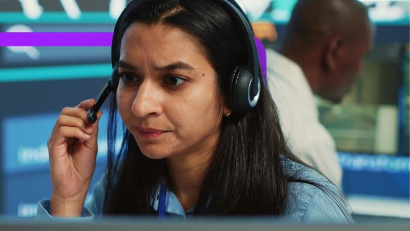 The image shows a close-up of a woman with dark hair wearing a headset with a microphone, concentrating intensely. She appears to be working in a control room or office with multiple screens visible in the background, and another person is partially visible behind her.
